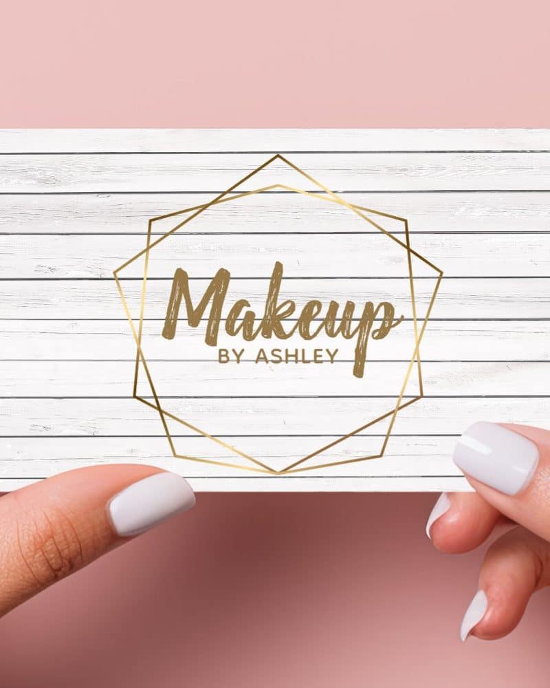 Two hands hold a business card with a white wood grain background. A gold geometric frame encloses the text Makeup By Ashley.