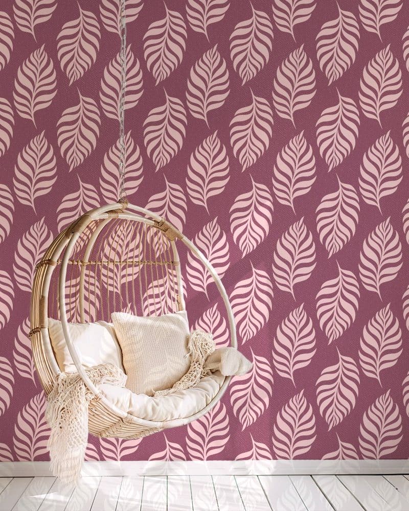 A hanging wicker chair with cushions sits in front of a wall with a pink leaf patterned wallpaper.