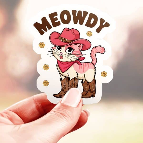 A Hand Holds A Sticker Of A Pink Cat Wearing A Cowboy Hat And Boots. The Words Meowdy Are Above The Cat.