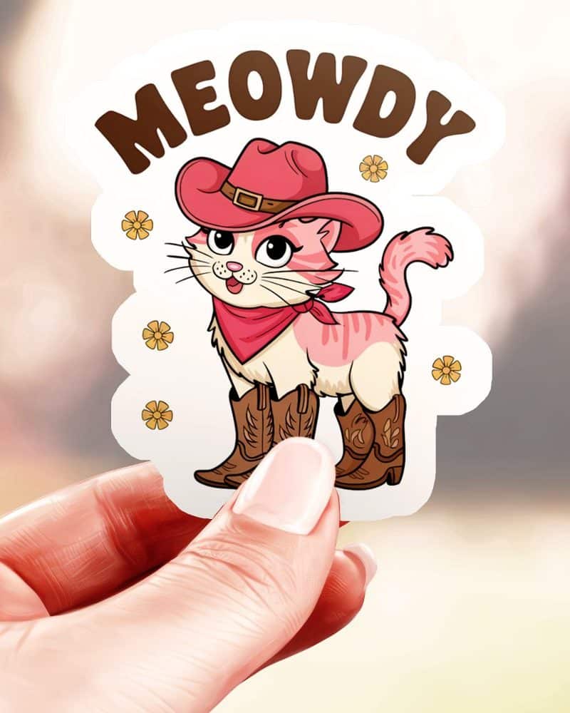 A hand holds a sticker of a pink cat wearing a cowboy hat and boots. The words MEOWDY are above the cat.