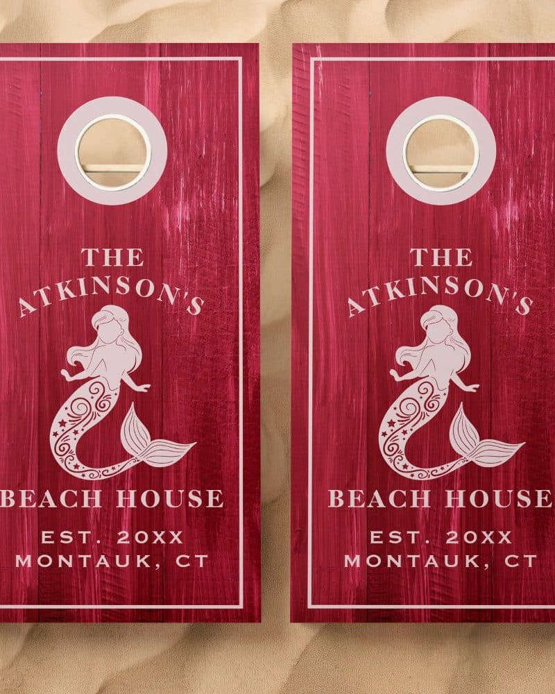 Pair of dark red wooden cornhole boards, each featuring a mermaid design, The Atkinsons Beach House text, and establishment year.