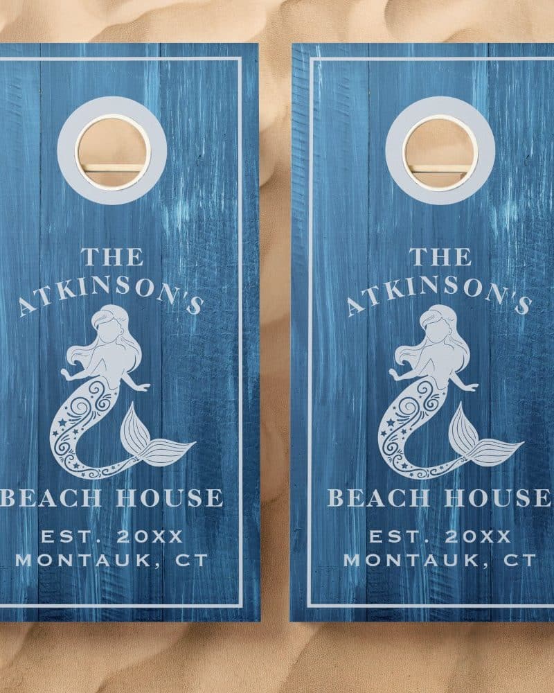 Two blue wooden cornhole boards, each featuring a mermaid design, the family name Atkinsons, Beach House, and establishment year.