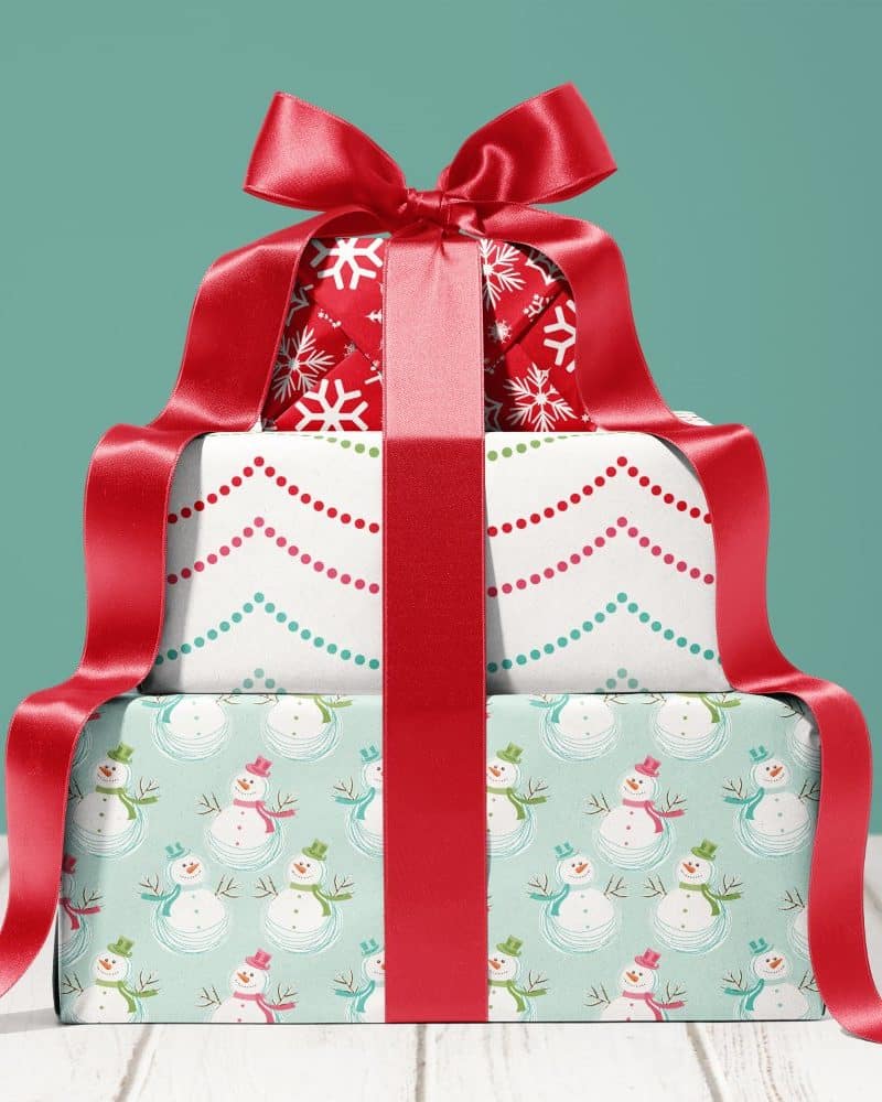 Three stacked Christmas presents tied with a large red ribbon. The top present has a snowflake design, the middle a dotted pattern, and the bottom has a snowman pattern.