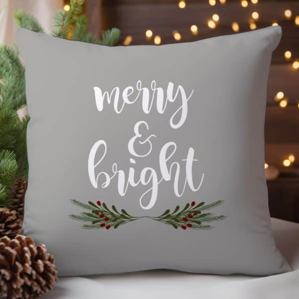 Gray Throw Pillow With White Script Lettering Reading Merry &Amp; Bright, Accented By Small Green Sprigs With Red Berries.