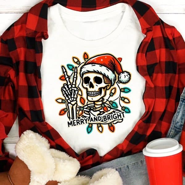 White Shirt Featuring A Skeleton Wearing A Santa Hat, Making A Peace Sign, Surrounded By Christmas Lights. The Text Merry And Bright Is Below.