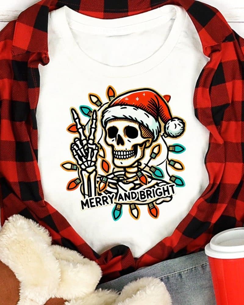 White shirt featuring a skeleton wearing a Santa hat, making a peace sign, surrounded by Christmas lights. The text Merry and Bright is below.