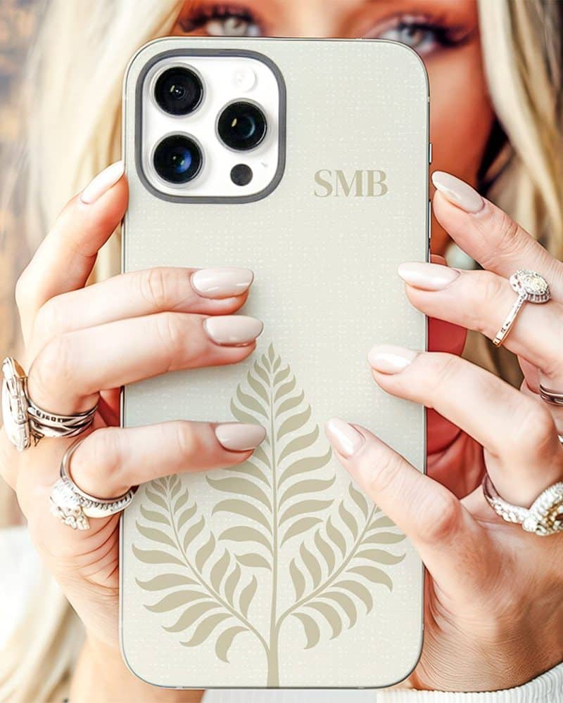 Close-up of a woman's hands holding a cream-colored iPhone case featuring a beige leaf design and initials.