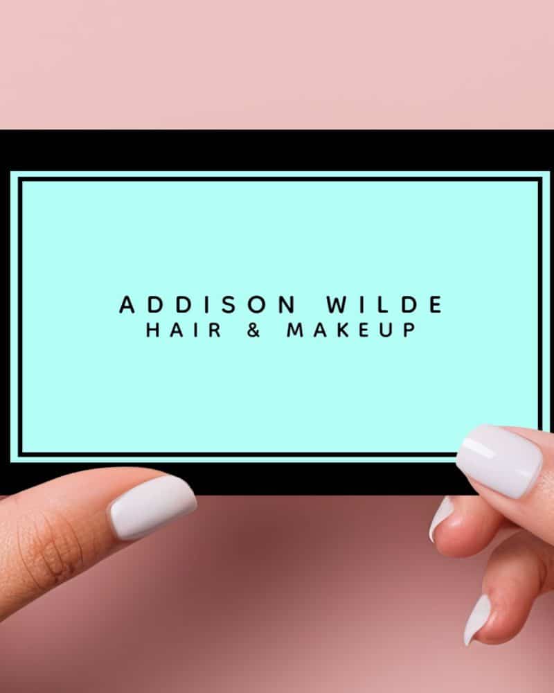 Two hands hold up a business card for Addison Wilde Hair & Makeup. The card has a light teal background with black text and border.
