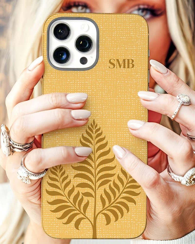 A woman's hands holding a mustard yellow iPhone case with a gold leaf design and monogram.