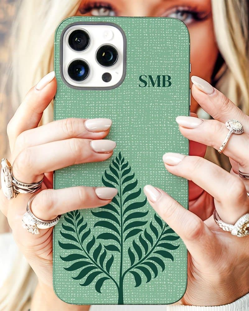 A woman's hands hold up a sage green iPhone case featuring a dark green fern design and personalized initials.