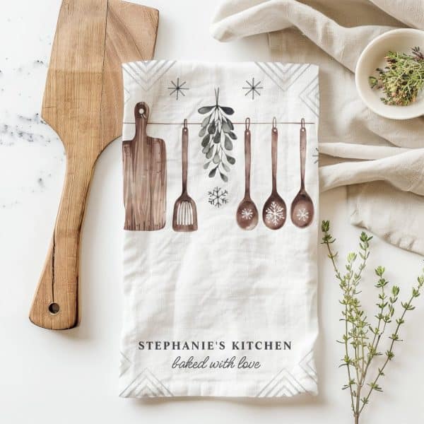 Kitchen Tea Towel With Watercolor Illustration Of Wooden Cooking Utensils Hanging With Mistletoe And Snowflakes. The Text Stephanie'S Kitchen Baked With Love Is At The Bottom.