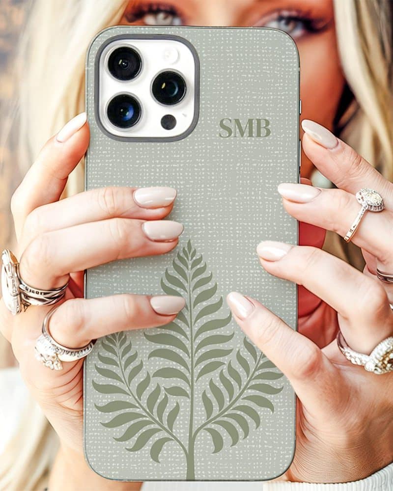 A woman's hands hold a light beige iPhone case featuring a fern design and initials SMB.