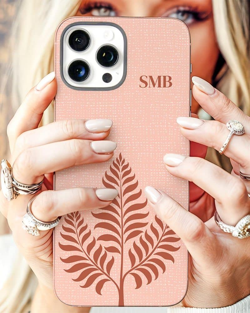 A woman's hands hold up a blush pink phone case with a fern design and initials SMB.