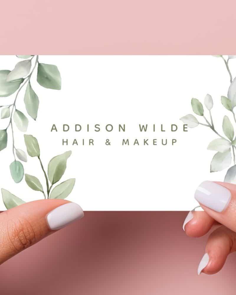 Two hands hold a business card featuring a watercolor design of green leaves and the text Addison Wilde Hair & Makeup.
