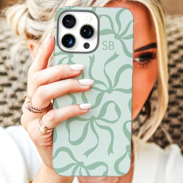 A Woman'S Hand Holds Up A Light Green Iphone Case Featuring A Repeating Pattern Of Bows. The Initials Sb Are Visible On The Case.