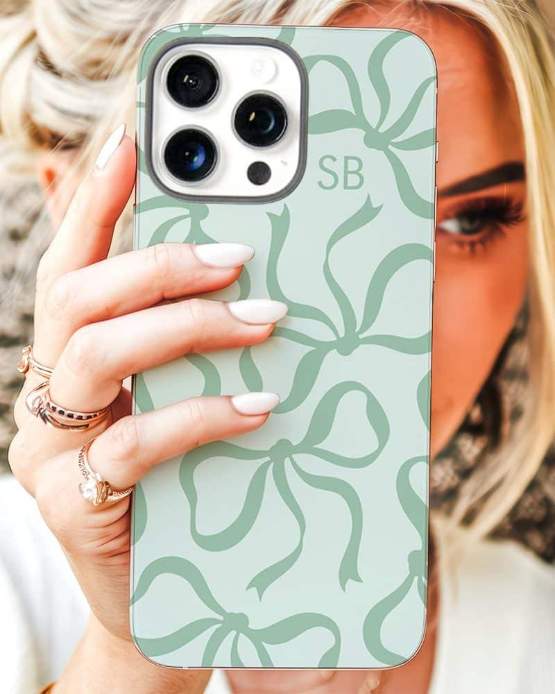 A woman's hand holds up a light green iPhone case featuring a repeating pattern of bows. The initials SB are visible on the case.