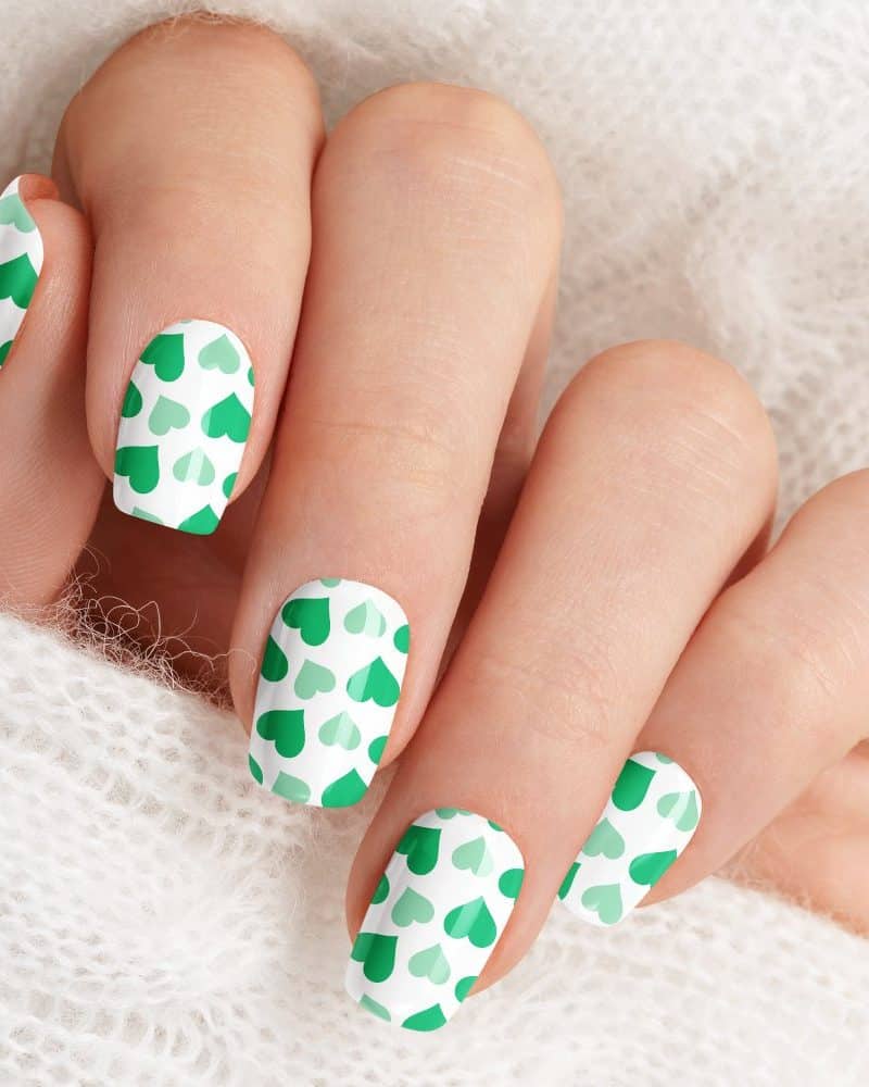 Closeup of a hand with fingernails painted with a pattern of small green hearts on a white background.