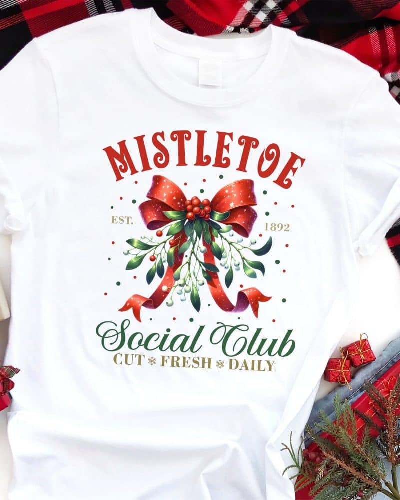 White t-shirt with a Christmas design featuring a mistletoe sprig tied with a red bow, text reading Mistletoe Social Club, and the year 1892.