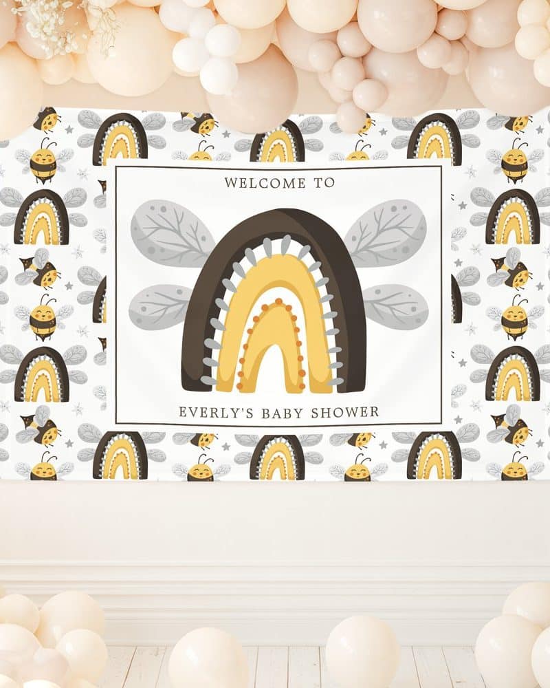 A baby shower banner featuring a large rainbow with bee wings and small bees and rainbows repeating on a white background. The banner also includes the text Welcome to Everly's Baby Shower.