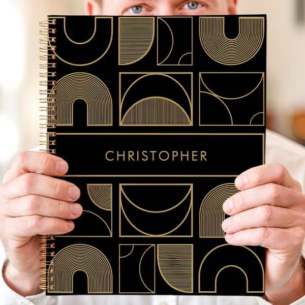 Person Holding A Black Notebook With Gold Geometric Designs And The Name Christopher.