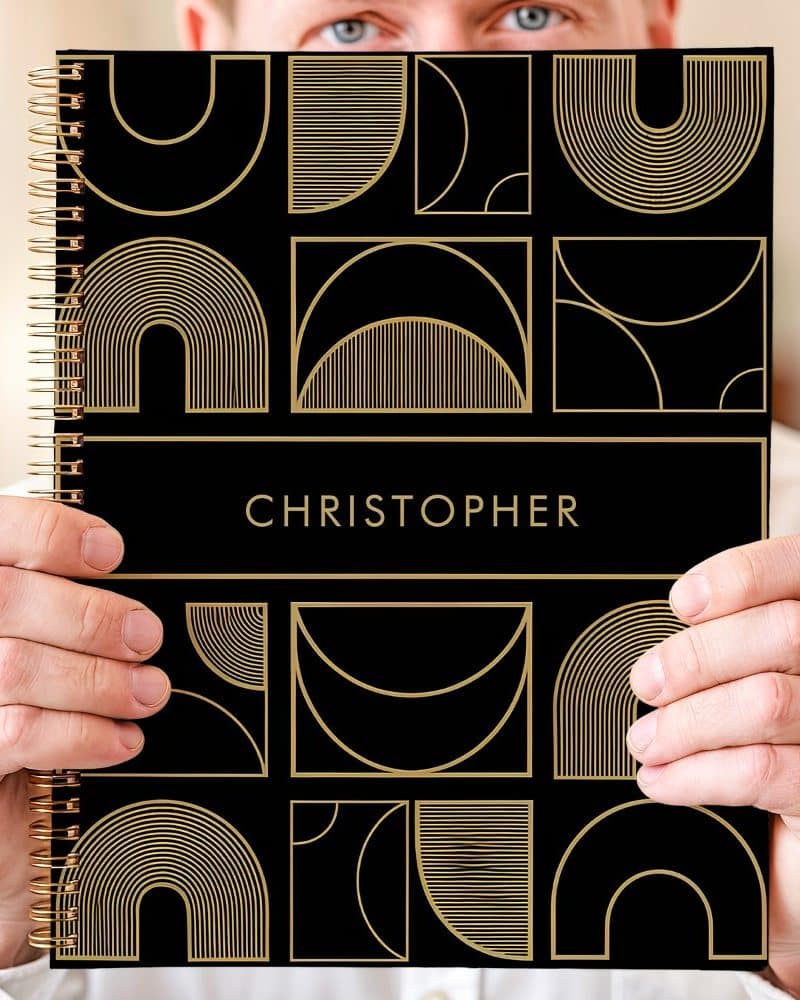 Person holding a black notebook with gold geometric designs and the name Christopher.