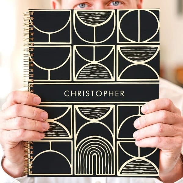 Person Holding A Black Spiral Notebook With A Gold Geometric Design And The Name Christopher On The Cover.