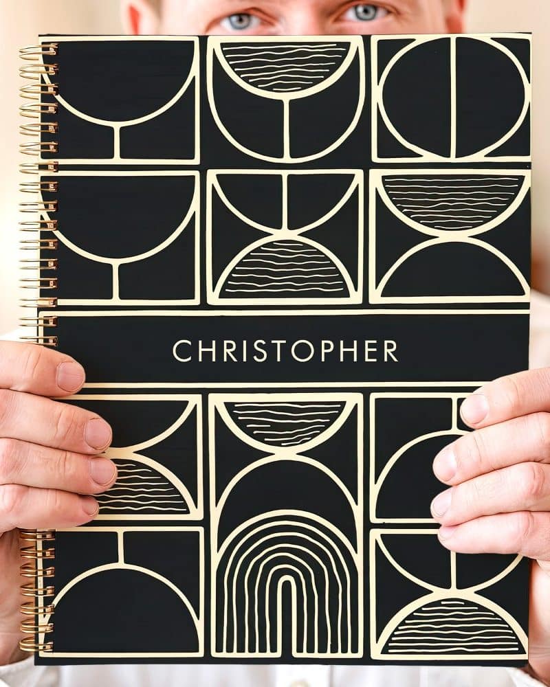 Person holding a black spiral notebook with a gold geometric design and the name Christopher on the cover.