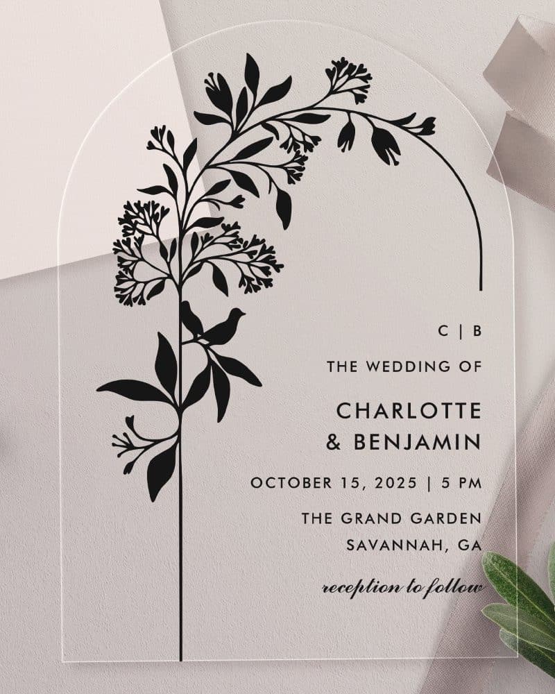 Arch-shaped clear acrylic wedding invitation with a black floral design and bird. The invitation features the couple's names and wedding details.