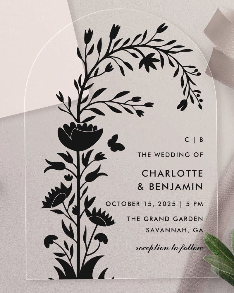 Arch-shaped clear acrylic wedding invitation with a black floral design and wedding details.