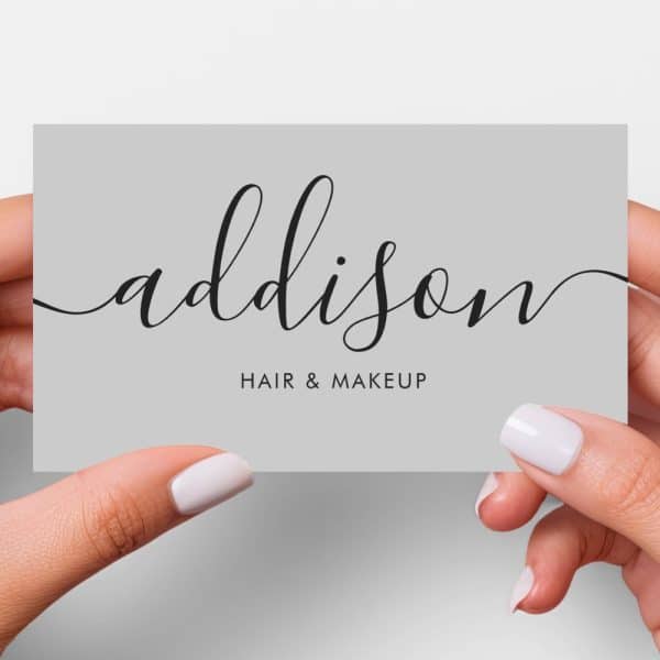 Two Hands Hold A Gray Business Card With Addison Hair &Amp; Makeup In Elegant Script.