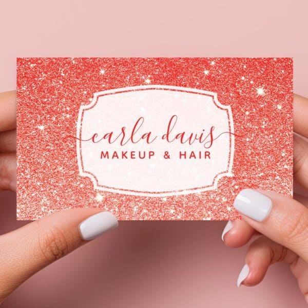 Two Hands Hold Up A Red Glitter Business Card That Says Carla Davis Makeup &Amp; Hair.