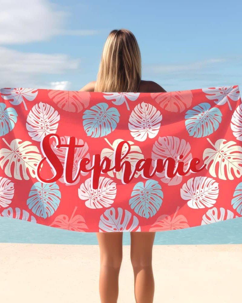 A woman holds a coral beach towel with a tropical leaf pattern and the name Stephanie printed on it.