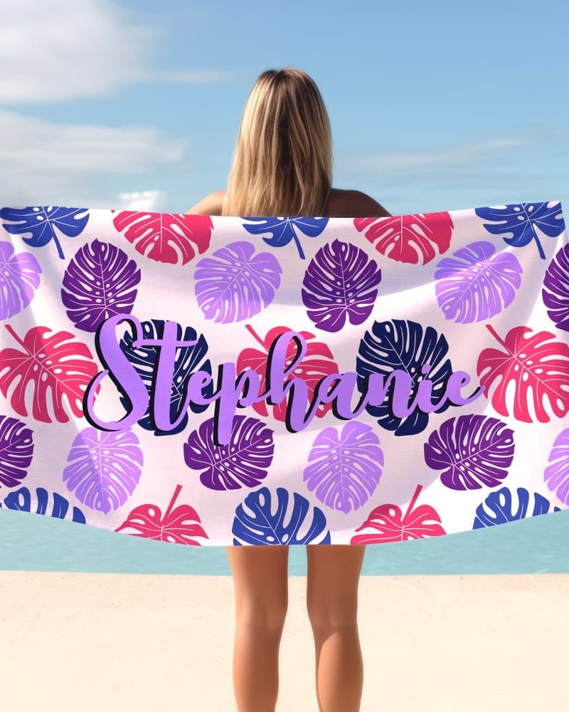 A woman holds a personalized beach towel featuring a tropical leaf pattern in shades of purple, pink, and blue. The name Stephanie is printed in purple script.