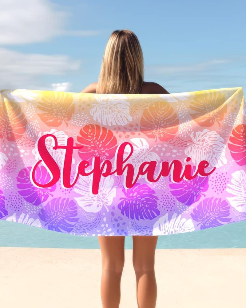 Woman holding a personalized beach towel with a tropical leaf pattern and her name, Stephanie, in pink lettering.
