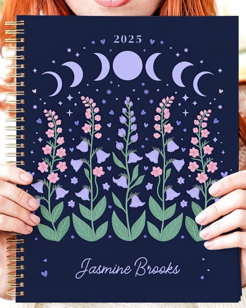 Person holding a 2025 spiral-bound planner featuring a dark blue cover with floral and moon phase illustrations. The name Jasmine Brooks is printed at the bottom.