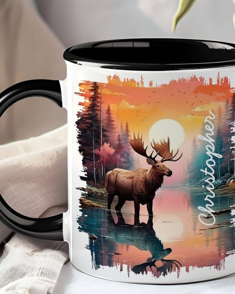 A black-handled mug features a painting of a moose standing in a calm lake at sunset, with the name Christopher printed on the side.