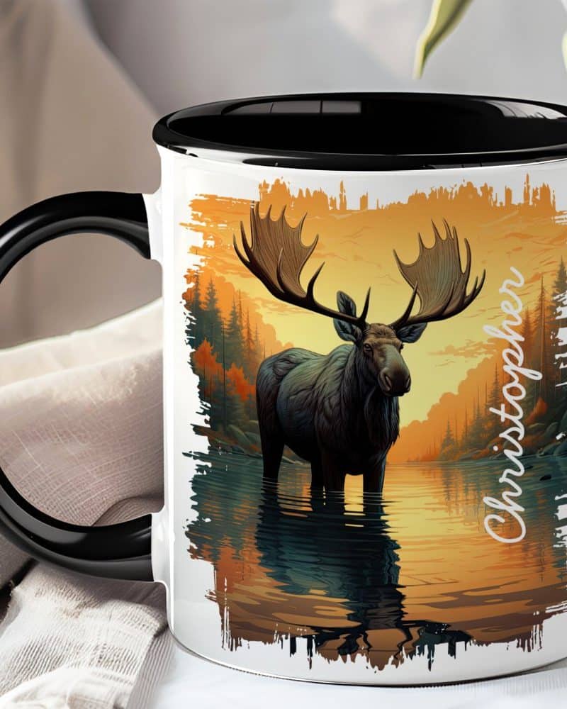 A coffee mug with a black handle and rim features a detailed illustration of a moose standing in a calm body of water at sunset. The name Christopher is printed on the mug.