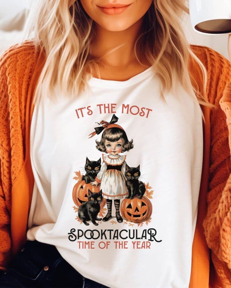 White shirt featuring a vintage illustration of a girl with three black cats and pumpkins. The text reads, It's the most Spooktacular Time of the Year.