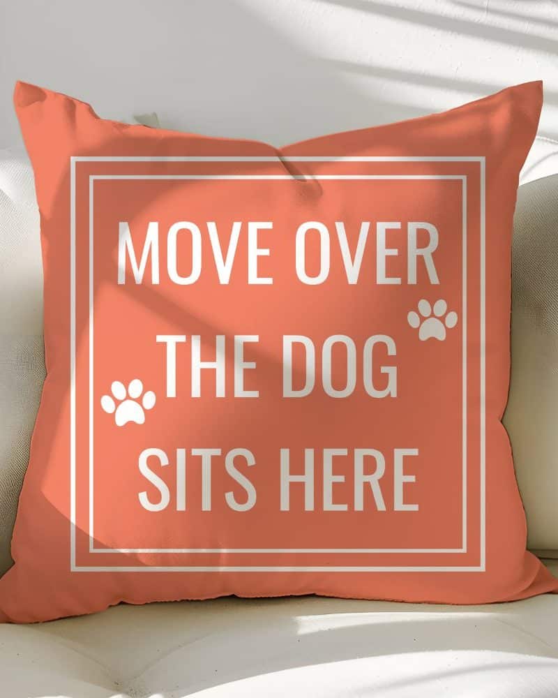 Coral-colored square pillow with the text Move Over The Dog Sits Here in white lettering and paw prints.