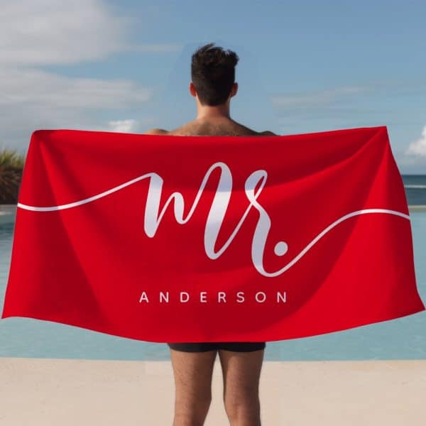 A Man Holds A Large Red Beach Towel Behind Him. The Towel Says &Quot;Mr. Anderson&Quot; In White Cursive Lettering.