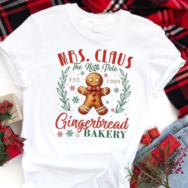 White T-Shirt With A Christmas Design Featuring A Gingerbread Man, Mrs. Claus'S Gingerbread Bakery Logo, And The Text &Quot;Est. 1989&Quot;.