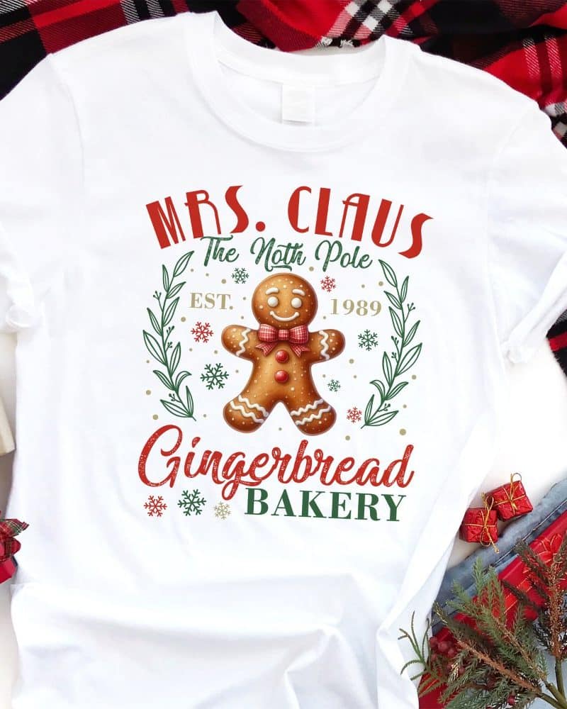 White t-shirt with a Christmas design featuring a gingerbread man, Mrs. Claus's Gingerbread Bakery logo, and the text "Est. 1989".