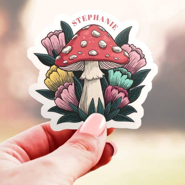 A Hand Holds A Sticker Featuring A Red Mushroom With White Dots Surrounded By Colorful Flowers. The Name Stephanie Is Printed Above The Mushroom.