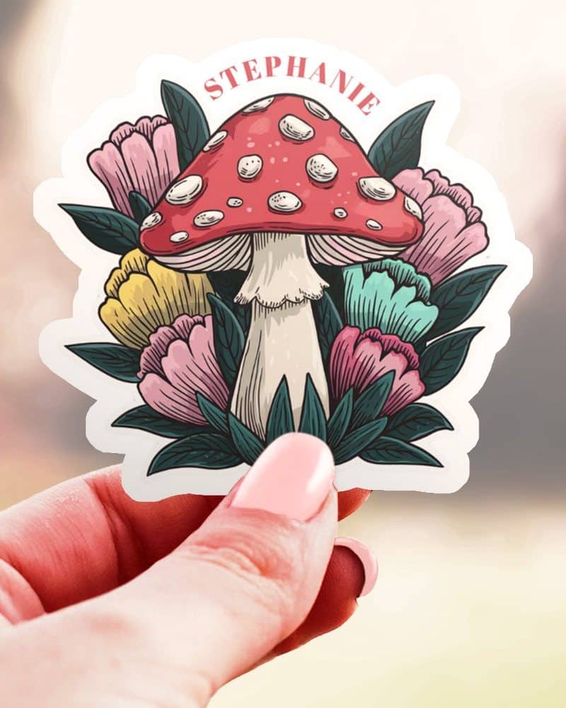 A hand holds a sticker featuring a red mushroom with white dots surrounded by colorful flowers. The name Stephanie is printed above the mushroom.