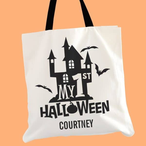 White Tote Bag With Black Halloween Design; A Haunted House Graphic, The Words My 1St Halloween, And A Name.