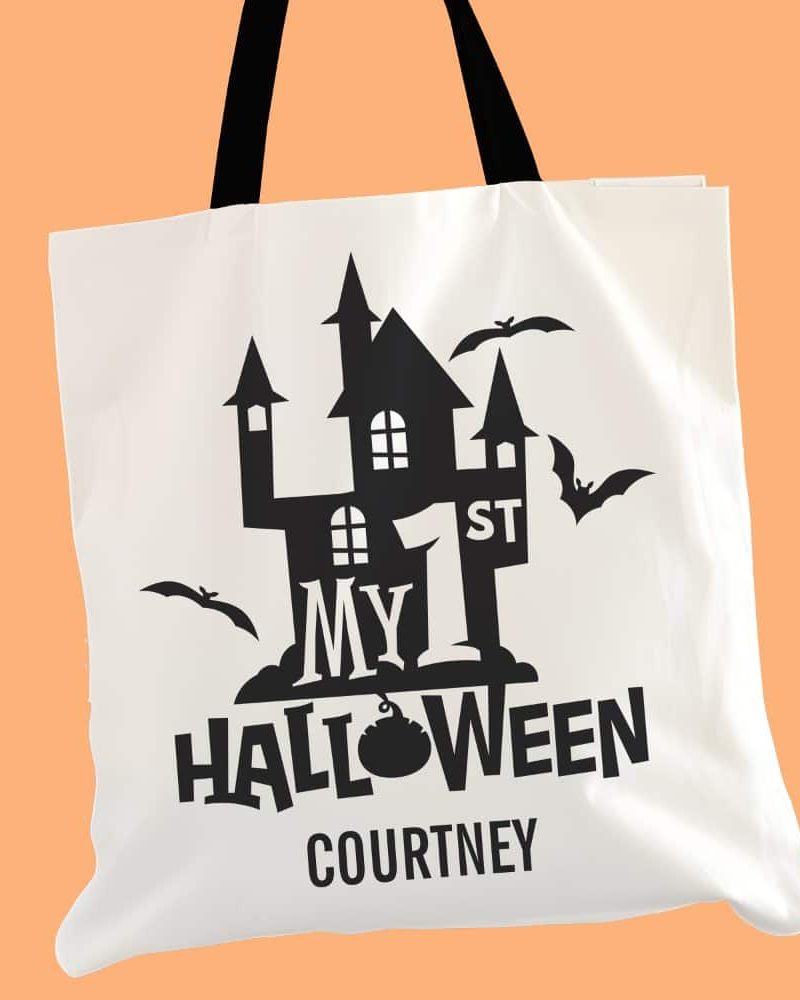 White tote bag with black Halloween design; a haunted house graphic, the words My 1st Halloween, and a name.