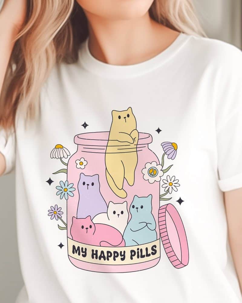 White t-shirt featuring a graphic of pastel colored cats inside a pink jar, surrounded by flowers, with the text My Happy Pills.