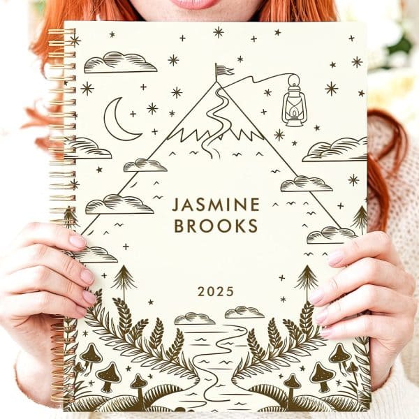 A Person Holds A Cream-Colored 2025 Planner Featuring A Mountain Scene With A Winding Path, A Lantern, And Mushrooms. The Name Jasmine Brooks Is Printed On The Cover.