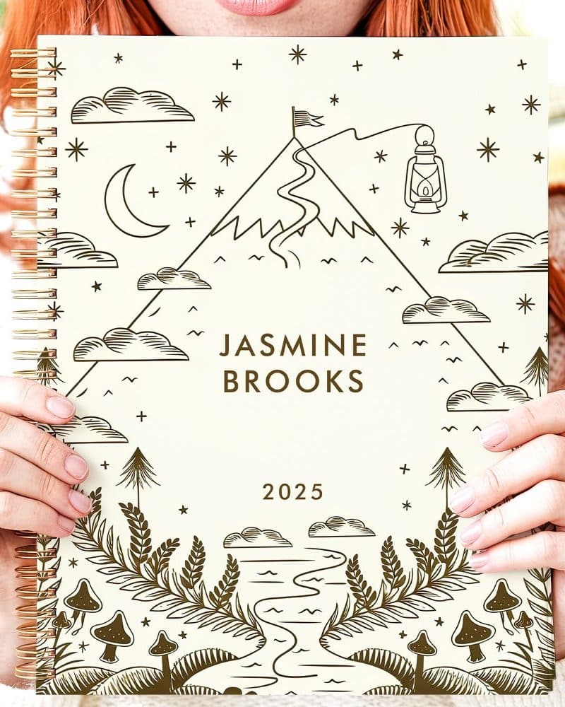 A person holds a cream-colored 2025 planner featuring a mountain scene with a winding path, a lantern, and mushrooms. The name Jasmine Brooks is printed on the cover.