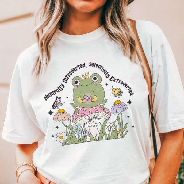 White T-Shirt Featuring A Cartoon Frog Wearing A Crown And Sitting On A Mushroom, Surrounded By Flowers And A Butterfly; Text Reads Naturally Introverted, Selectively Extroverted.